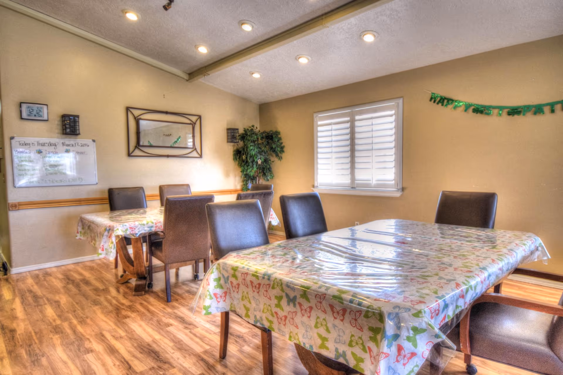 A dining room with two tables covered in butterfly-patterned tablecloths and clear plastic covers. Each table is surrounded by brown chairs. The room has wooden flooring, beige walls, a window with white shutters, a wall mirror, a whiteboard with the day's menu, and a green plant in the corner. A green banner on the wall reads 'Happy St. Patrick's Day.'