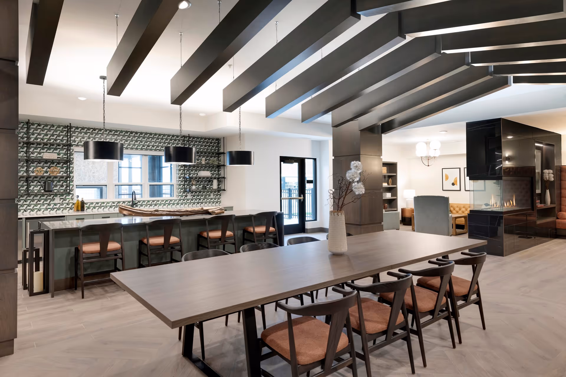 Modern senior living facility common area with a long wooden dining table surrounded by chairs with brown cushions. Behind the table is a kitchen island with four bar stools and three black pendant lights hanging above. The kitchen backsplash features a green and white geometric tile pattern. To the right, there is a black glass fireplace with seating areas including a tufted sofa and armchairs. The ceiling has exposed dark wooden beams and the floor is light wood.