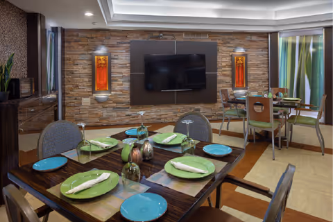 A dining area with a wooden table set for six people with green and blue plates, napkins, and glasses. The room features a stone accent wall with a mounted flat-screen TV and two decorative wall sconces. Additional tables and chairs are visible in the background, along with large windows with curtains.