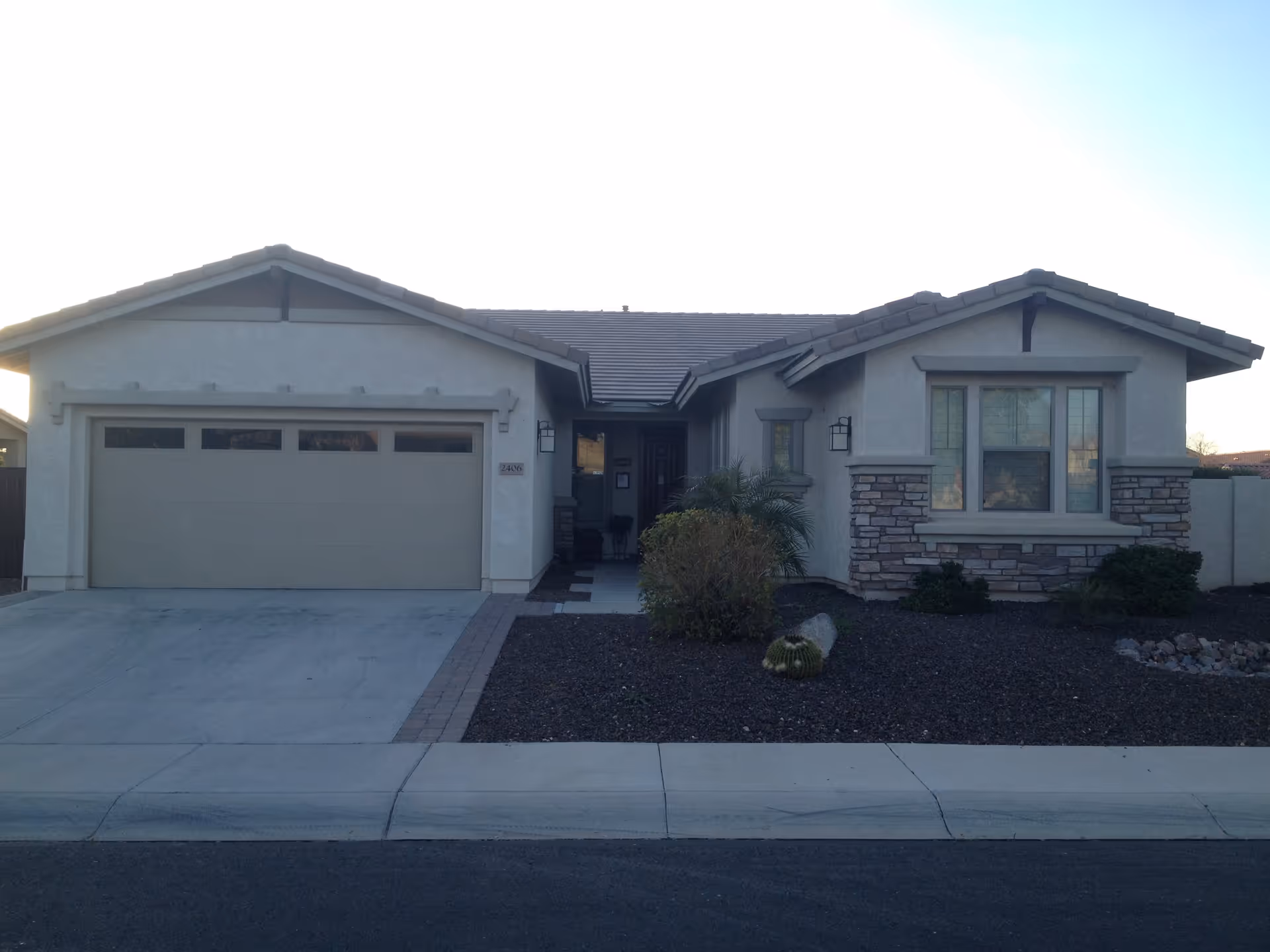 Front exterior view of a single-story house with a two-car garage, stone and stucco facade, a small landscaped area with bushes and a cactus, and a paved driveway.