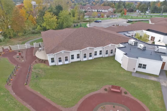 Aerial view of a single-story assisted living building with a red roof, surrounding lawn, curved walking paths and a bench.