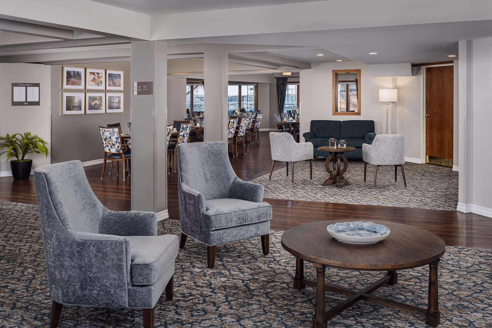 A spacious senior living community common area featuring two gray upholstered armchairs and a round wooden coffee table with a decorative bowl in the foreground. In the background, there is a seating area with a dark green sofa, two light gray chairs, a small round table, and a floor lamp. Further back, a dining area with multiple tables and chairs with floral upholstery is visible. The room has a patterned carpet, wooden flooring, neutral-colored walls, framed artwork, and large windows letting in natural light.