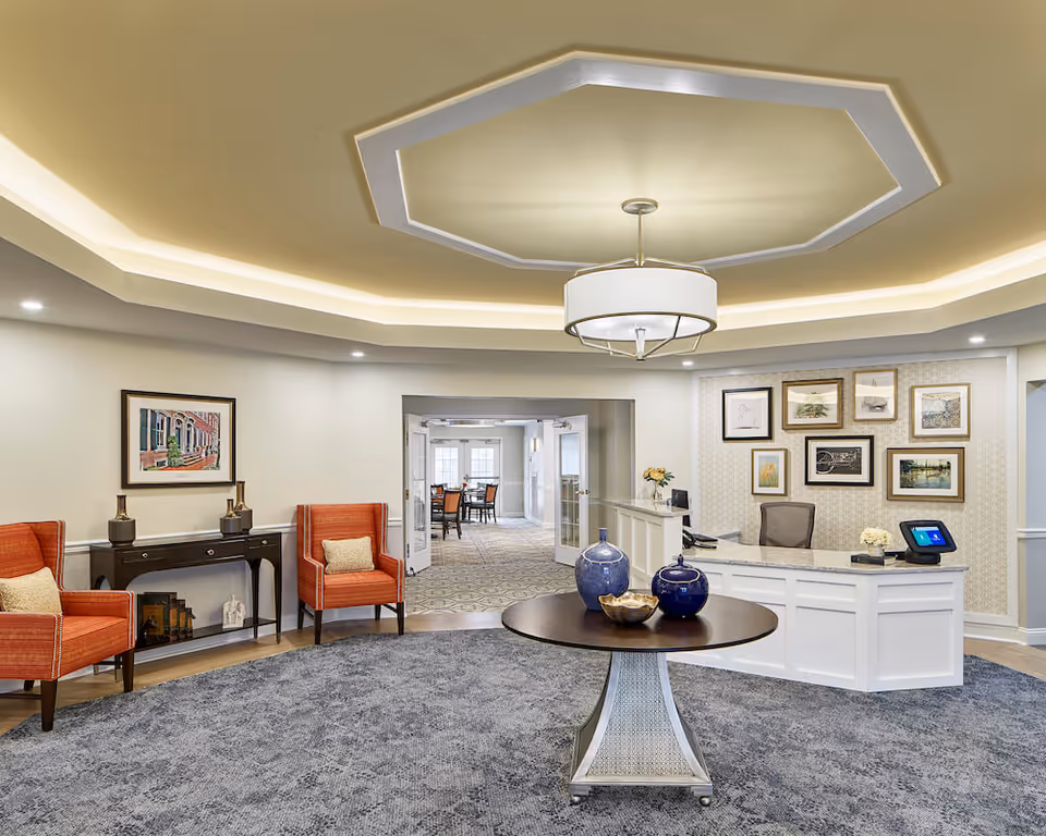 A bright and welcoming reception area in a senior living facility with a round table in the center holding decorative blue vases and a bowl. To the right is a white reception desk with a computer and chair, and behind it is a wall adorned with framed artwork. On the left side, there are two orange armchairs with beige cushions and a black console table with decorative items and a framed picture above it. The ceiling features recessed lighting and a modern chandelier. Through an open doorway, a dining area with tables and chairs is visible.
