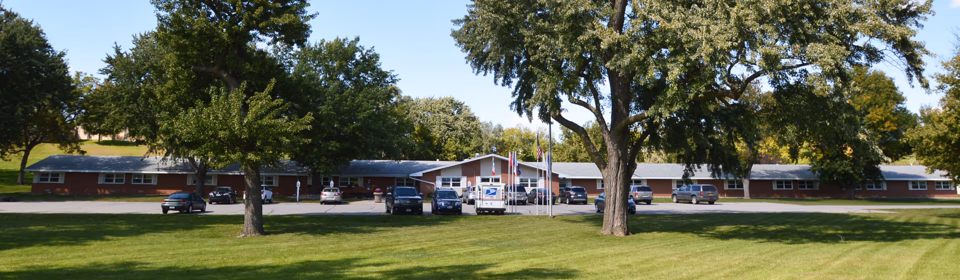 Wide exterior view of a single-story brick building with a parking lot in front, several cars parked, and multiple trees on a grassy lawn under a clear blue sky.