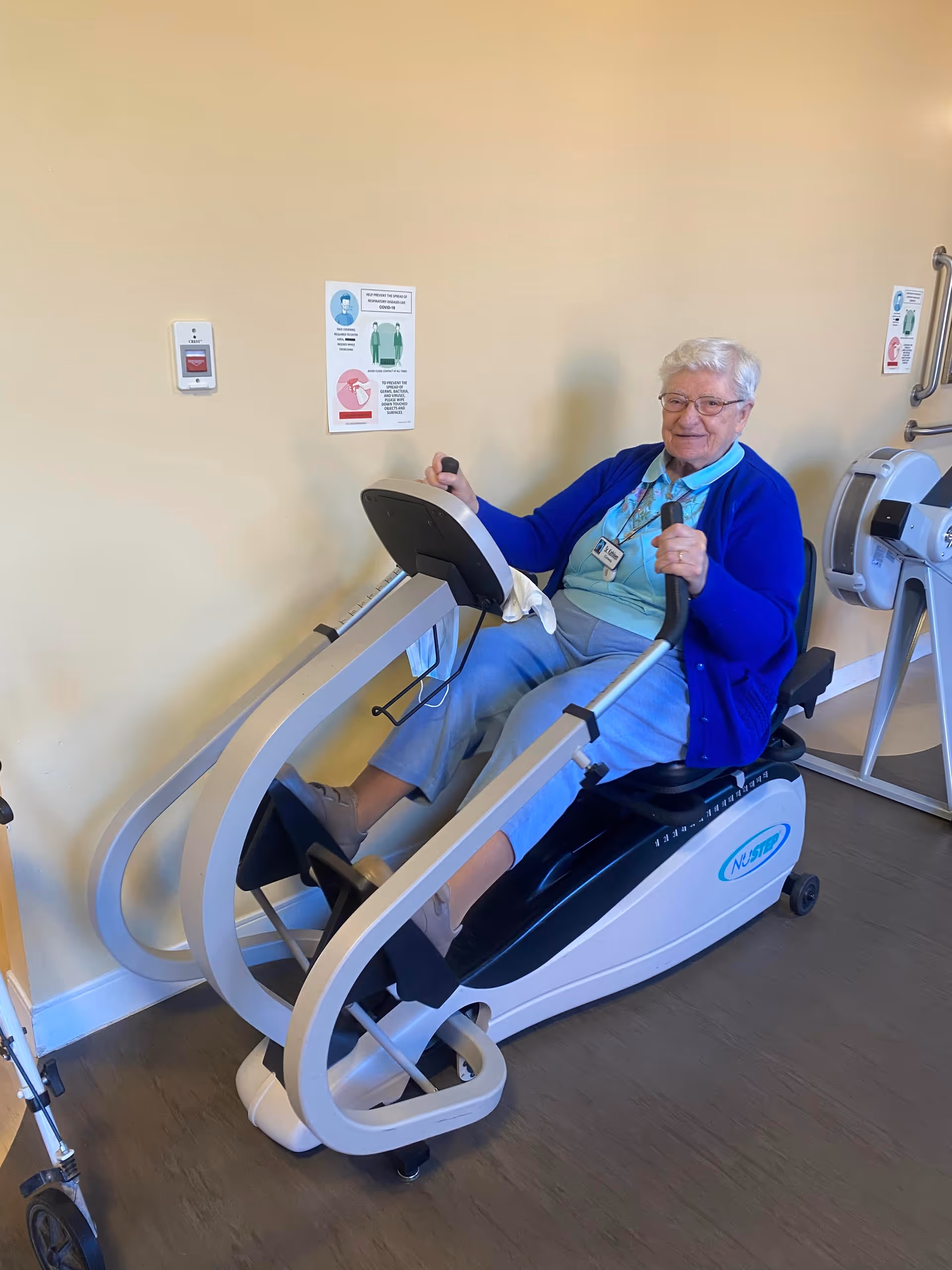 An elderly woman wearing glasses, a blue cardigan, and light blue pants is seated on a NuStep exercise machine in a room with beige walls. She is holding the machine's handles and smiling. There are safety and hygiene instruction signs on the wall behind her.