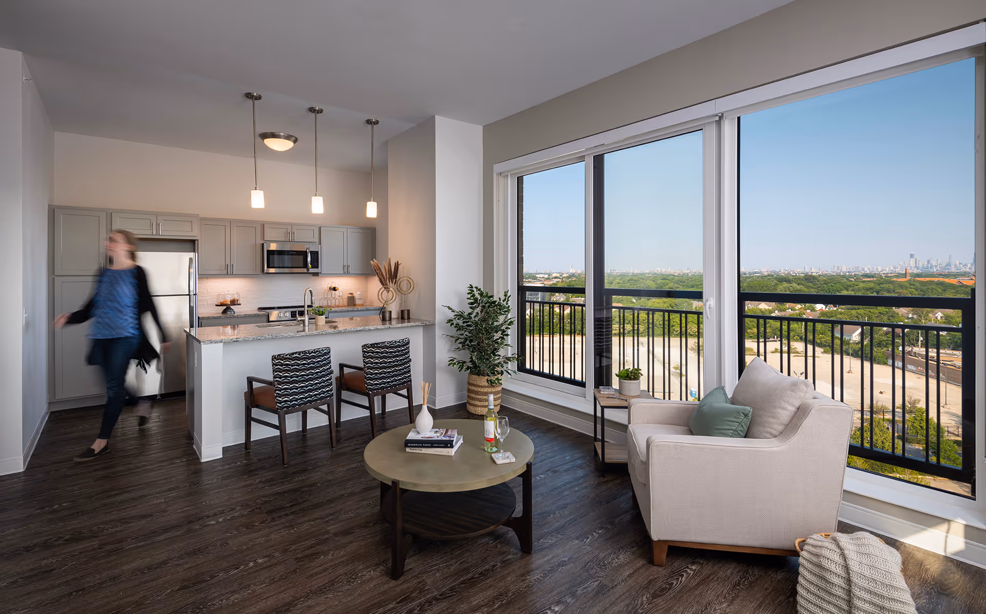 Open-plan living room and kitchen with large windows and a balcony overlooking a green landscape, an armchair and coffee table, and a person walking near the kitchen.