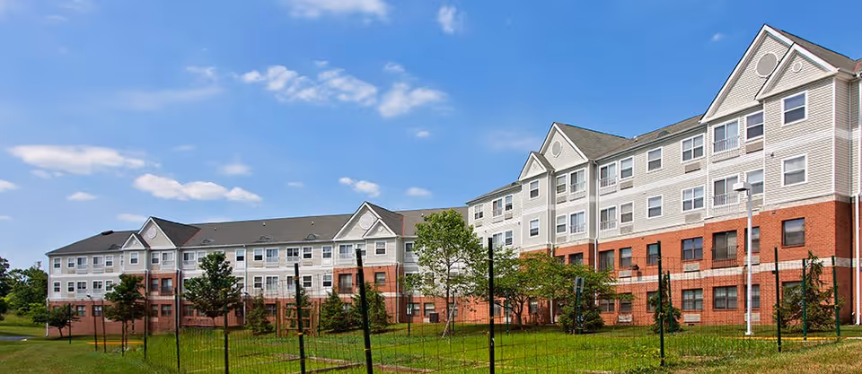Exterior view of a large multi-story senior living facility building with a combination of brick and light-colored siding under a blue sky with scattered clouds. The building is surrounded by green grass and some young trees, enclosed by a black metal fence.