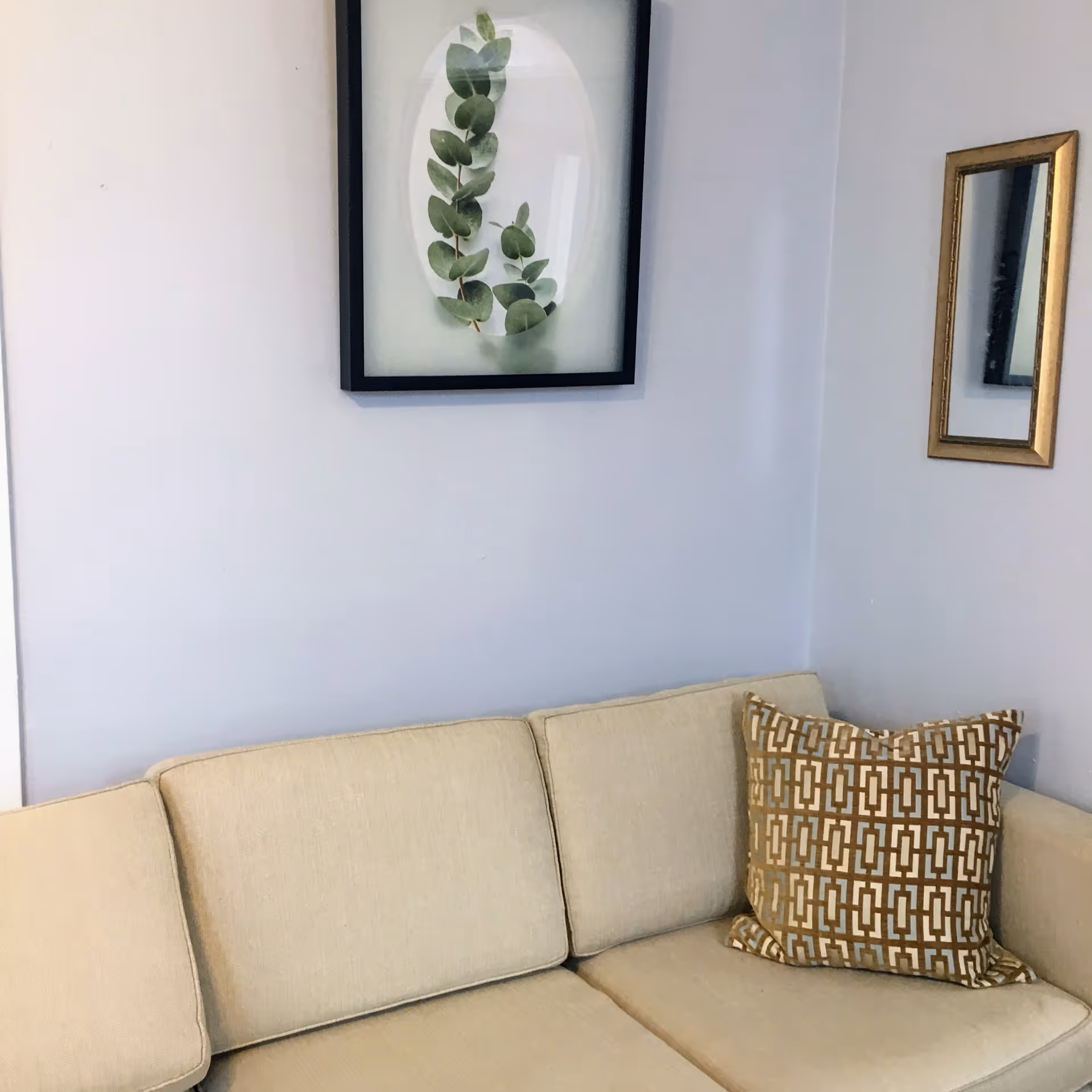 Corner of a living room with a beige sofa, a patterned throw pillow, and framed artwork and mirror on light-colored walls.