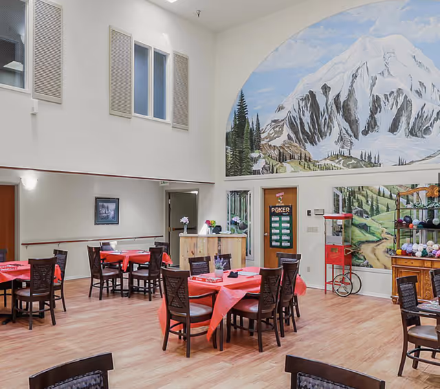 Spacious assisted living dining area with multiple round tables covered in red tablecloths, wooden chairs, a popcorn machine, and a large mountain mural on the wall.