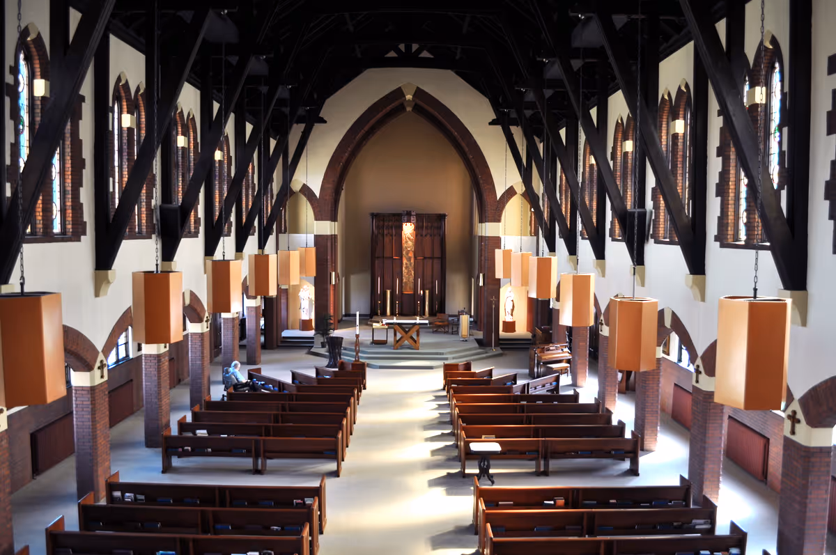 Interior view of a chapel with wooden pews arranged in rows facing an altar. The chapel features high vaulted ceilings with dark wooden beams, arched windows with stained glass, and hanging rectangular light fixtures. A person is seated on one of the pews near the front.