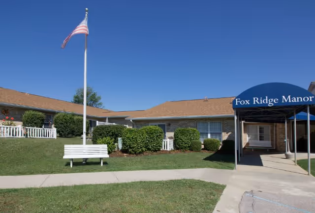 Exterior view of Fox Ridge Manor, a single-story brick building with a brown roof. There is a blue canopy over the entrance with the facility name on it. A white bench and an American flag on a flagpole are visible on the grassy area in front of the building, along with trimmed bushes and a clear blue sky.