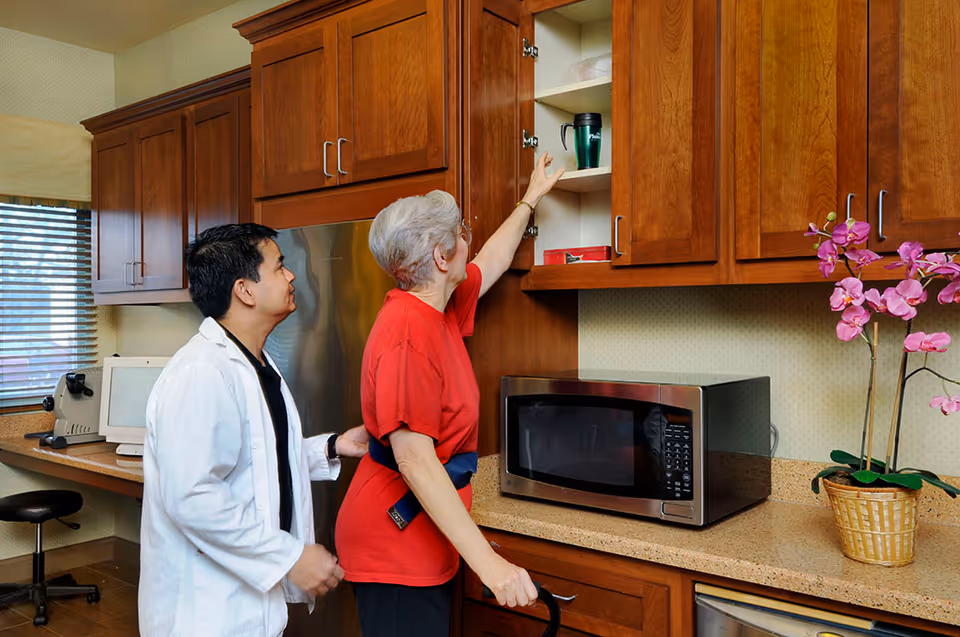 An elderly woman reaching for a cup in a kitchen cabinet with the assistance of a healthcare worker wearing a white coat. The kitchen has wooden cabinets, a microwave on the counter, and a potted orchid flower.