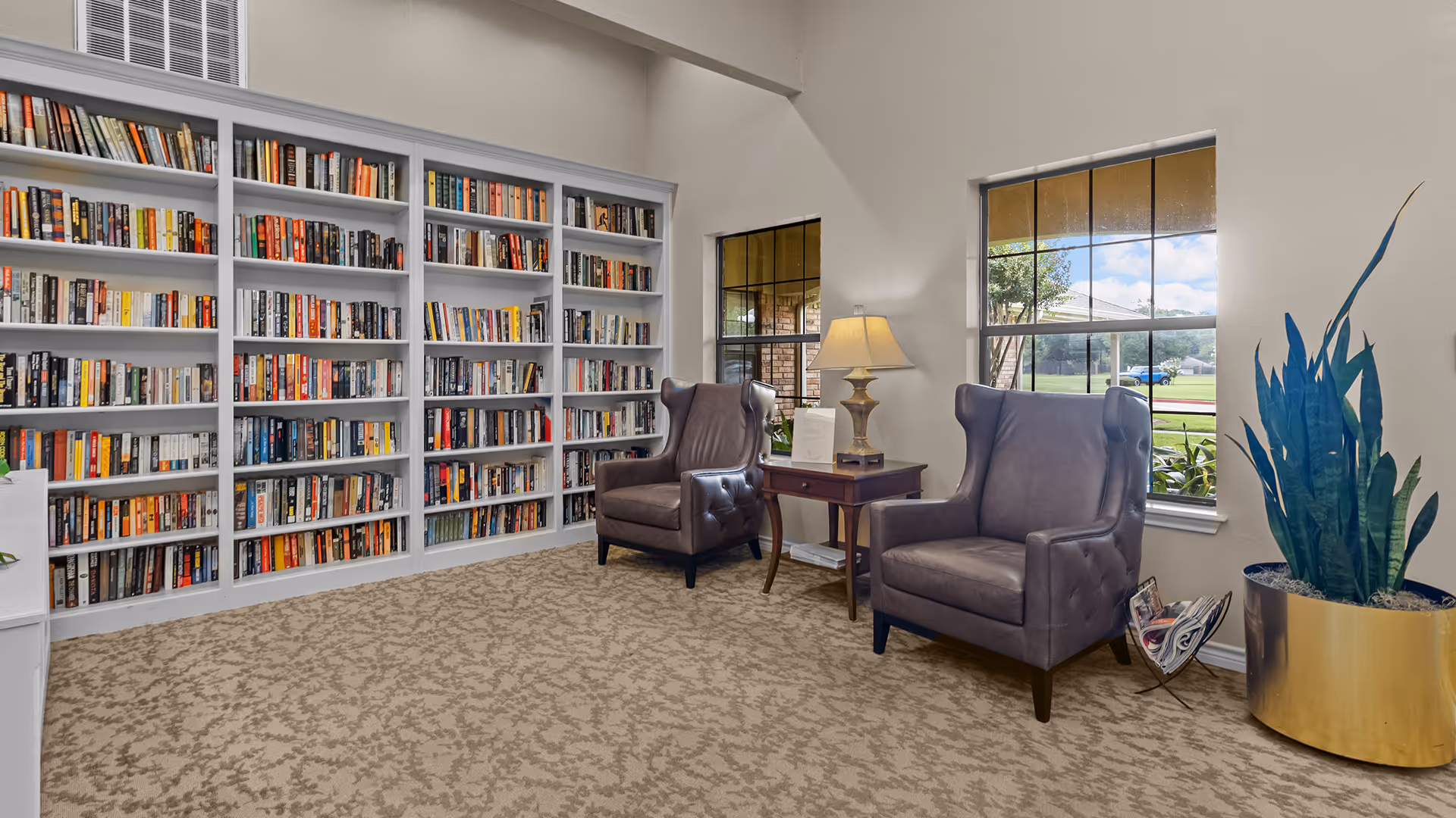 A cozy reading nook in a senior living facility featuring two brown leather armchairs separated by a wooden side table with a lamp. Behind the chairs is a large built-in bookshelf filled with books. Two windows allow natural light to brighten the room, and a large potted plant is placed near one window.