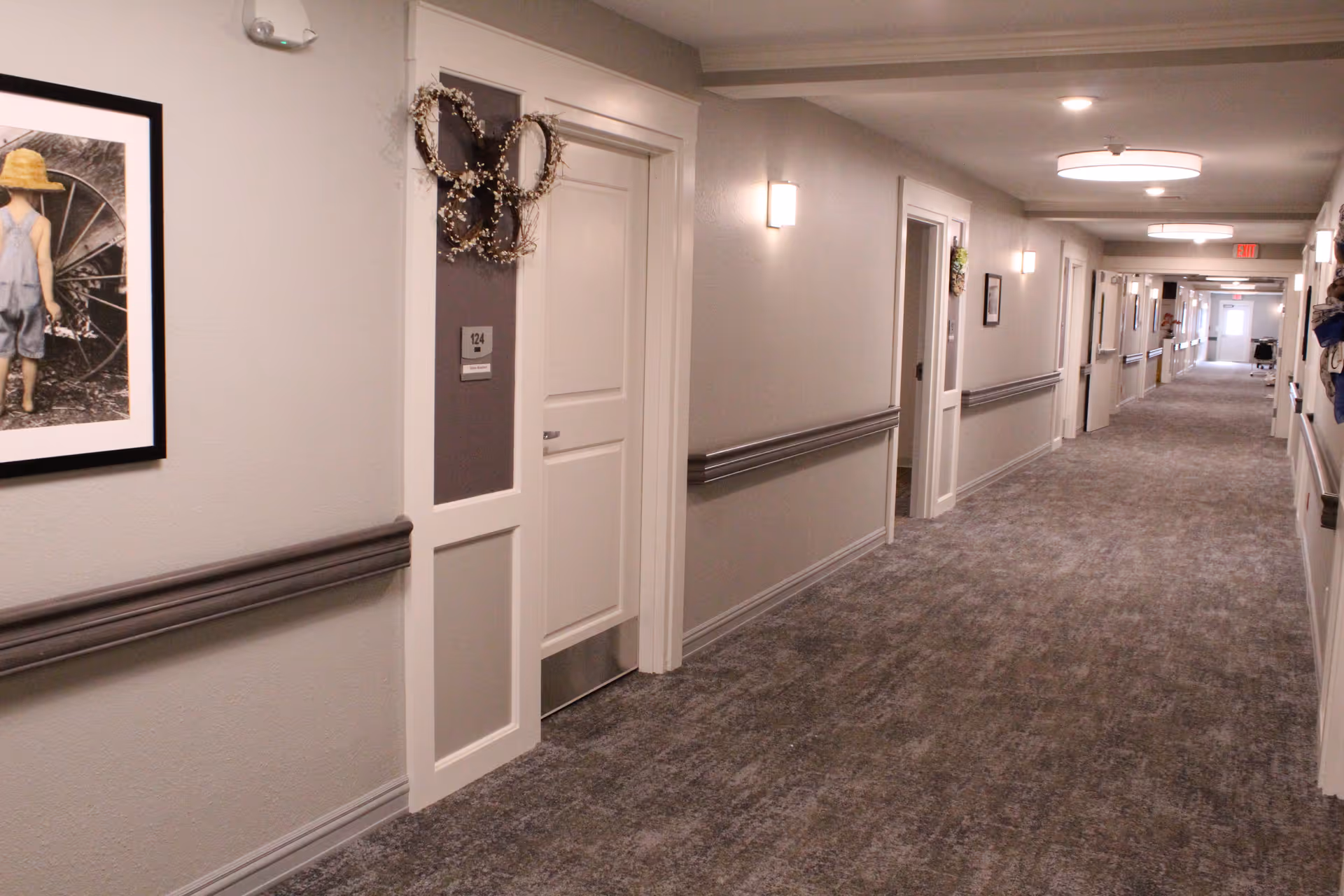Carpeted interior hallway of a senior living facility with numbered doors, handrails, and overhead lights.