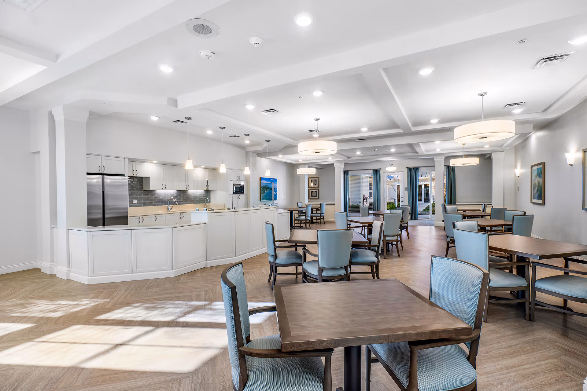 A bright and spacious dining room with multiple wooden tables and blue cushioned chairs. The room features a modern kitchen area with white cabinetry, stainless steel appliances, and pendant lighting. Large windows and glass doors allow natural light to fill the space, creating an inviting atmosphere.