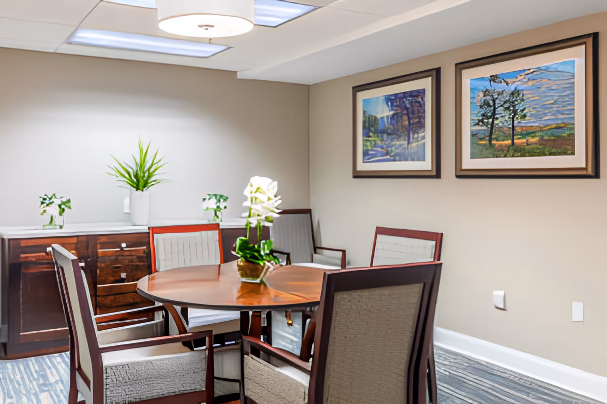 Small dining/meeting room with a round wooden table, upholstered chairs, potted plants, and framed artwork on the walls.