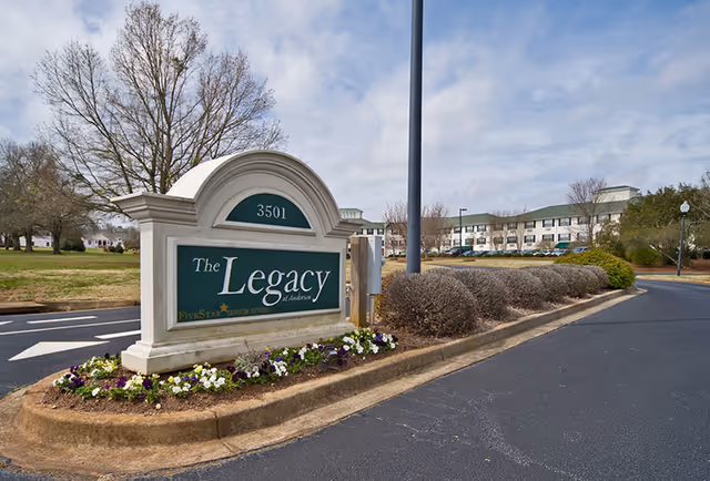 Entrance sign for The Legacy of Anderson senior living facility with the building and parking lot visible in the background under a partly cloudy sky.