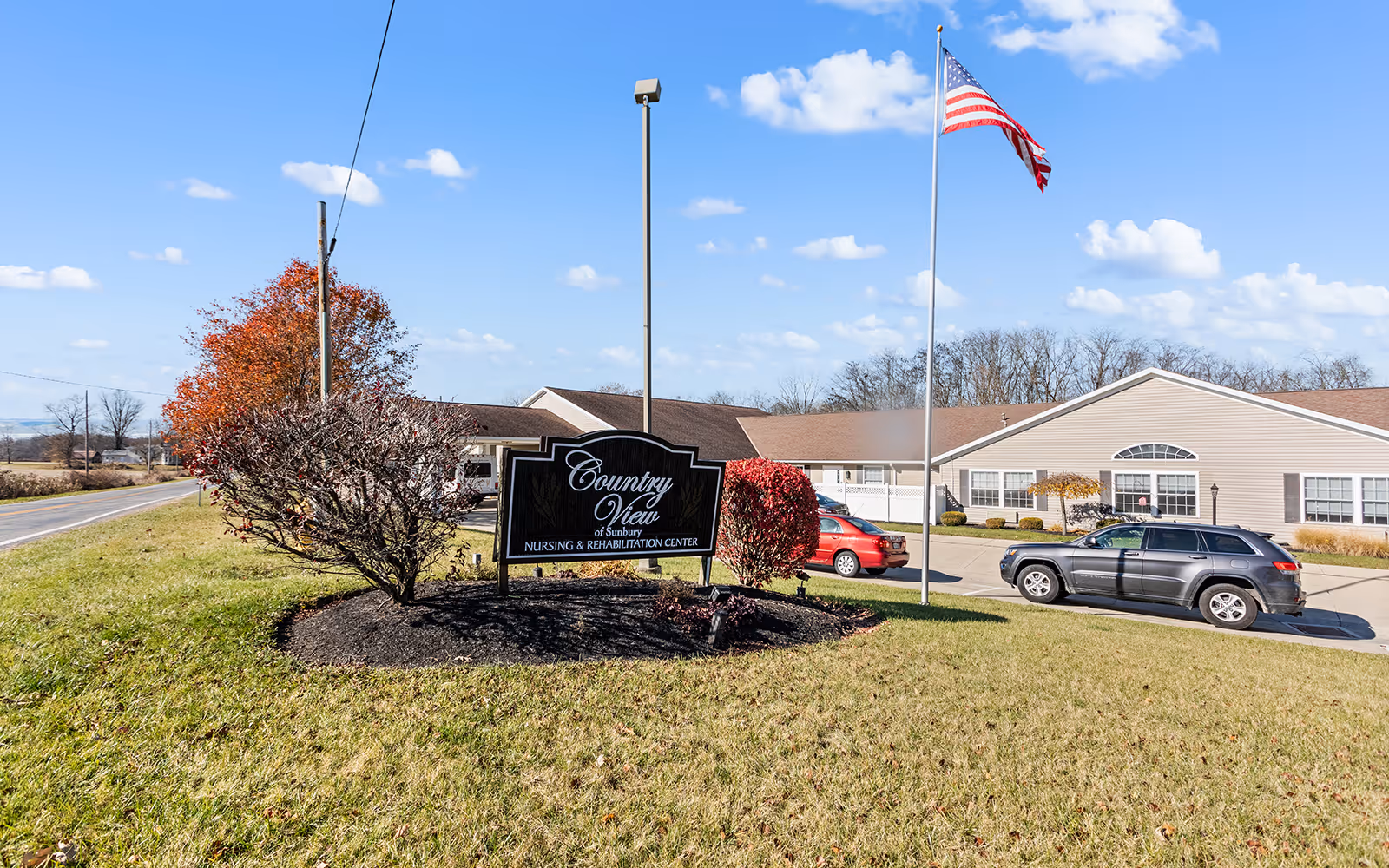Exterior view of Country View of Sunbury Nursing & Rehabilitation Center with a sign in the foreground, an American flag on a flagpole, two parked cars, and a clear blue sky with some clouds.