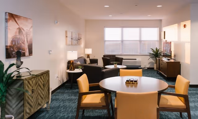 A well-lit living room area with a round table surrounded by four yellow cushioned chairs in the foreground. In the background, there are two dark armchairs and a gray sofa with side tables and lamps. A large window with blinds lets in natural light, and a flat-screen TV is placed on a wooden console against the wall. The room is decorated with plants and wall art.