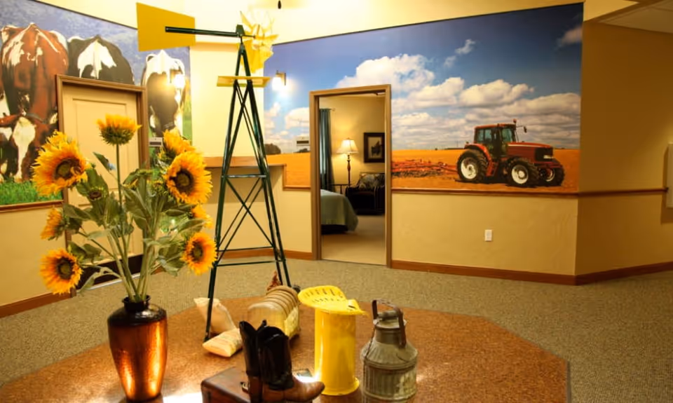 Interior room with a rustic farm theme featuring a large mural of cows and a tractor in a field. In the foreground, there is a table with a vase of sunflowers, a small decorative windmill, a yellow metal stool, cowboy boots, and a vintage milk can. A doorway leads to a bedroom with a lamp and bed visible.
