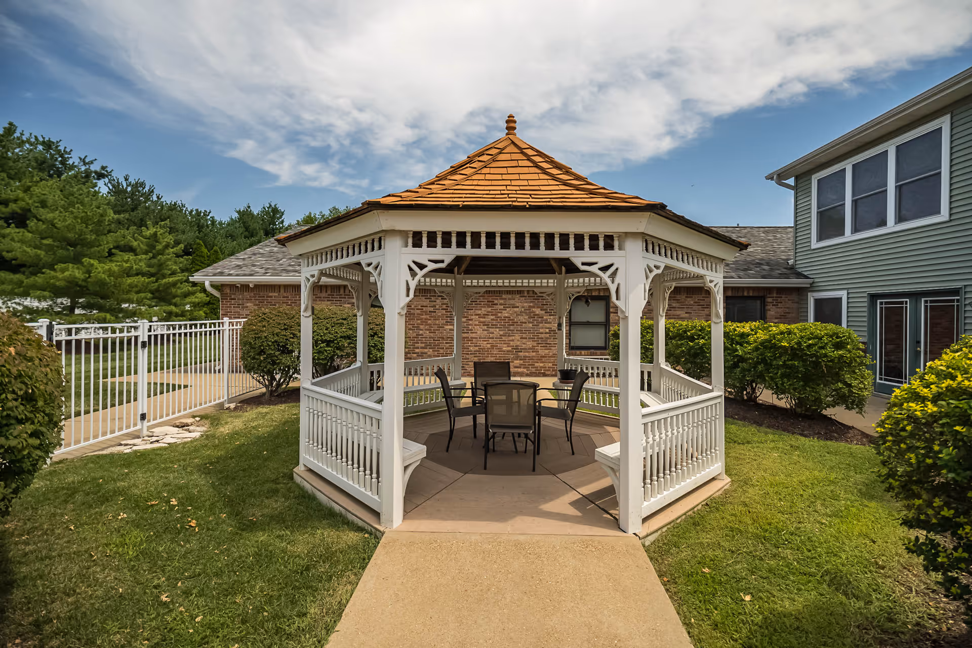A white wooden gazebo with a brown shingled roof situated on a concrete path in a grassy outdoor area. Inside the gazebo, there is a round table with four chairs. Surrounding the gazebo are bushes, a white metal fence, and parts of a brick and green siding building under a partly cloudy sky.