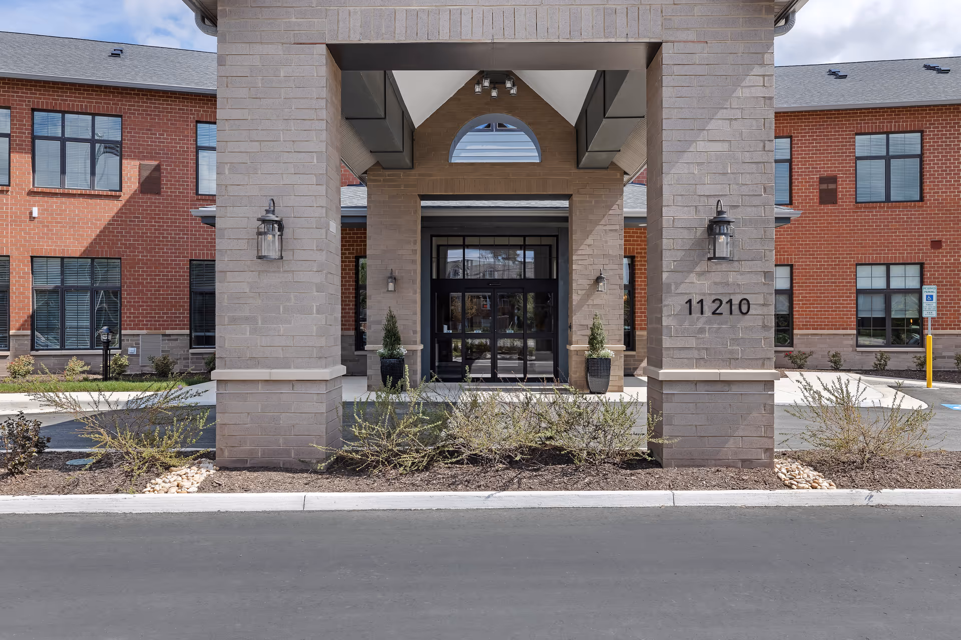 Front entrance of a senior living facility with brick and beige stone exterior, large pillars, glass double doors, and the number 11210 displayed on one pillar. There are small shrubs and plants in front of the entrance and a paved driveway.