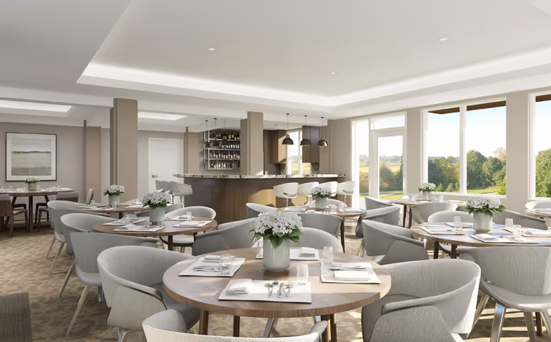 Bright and spacious dining room with multiple round wooden tables set with white napkins, silverware, and glasses. Each table has a vase with white flowers. Comfortable gray upholstered chairs surround the tables. In the background, there is a bar area with high stools and hanging pendant lights. Large windows let in natural light and offer a view of green trees and open landscape outside.