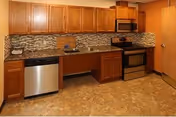 A kitchen area with wooden cabinets, a stainless steel dishwasher, a stainless steel oven with a stove, a microwave above the stove, a sink, and a tiled backsplash. The floor has a brownish tile pattern and the walls are painted in a warm orange tone.