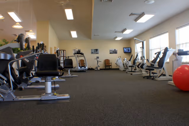 A spacious fitness room with various exercise machines including stationary bikes, elliptical trainers, and weight machines. The room has carpeted flooring, large windows letting in natural light, and a red exercise ball on the right side.