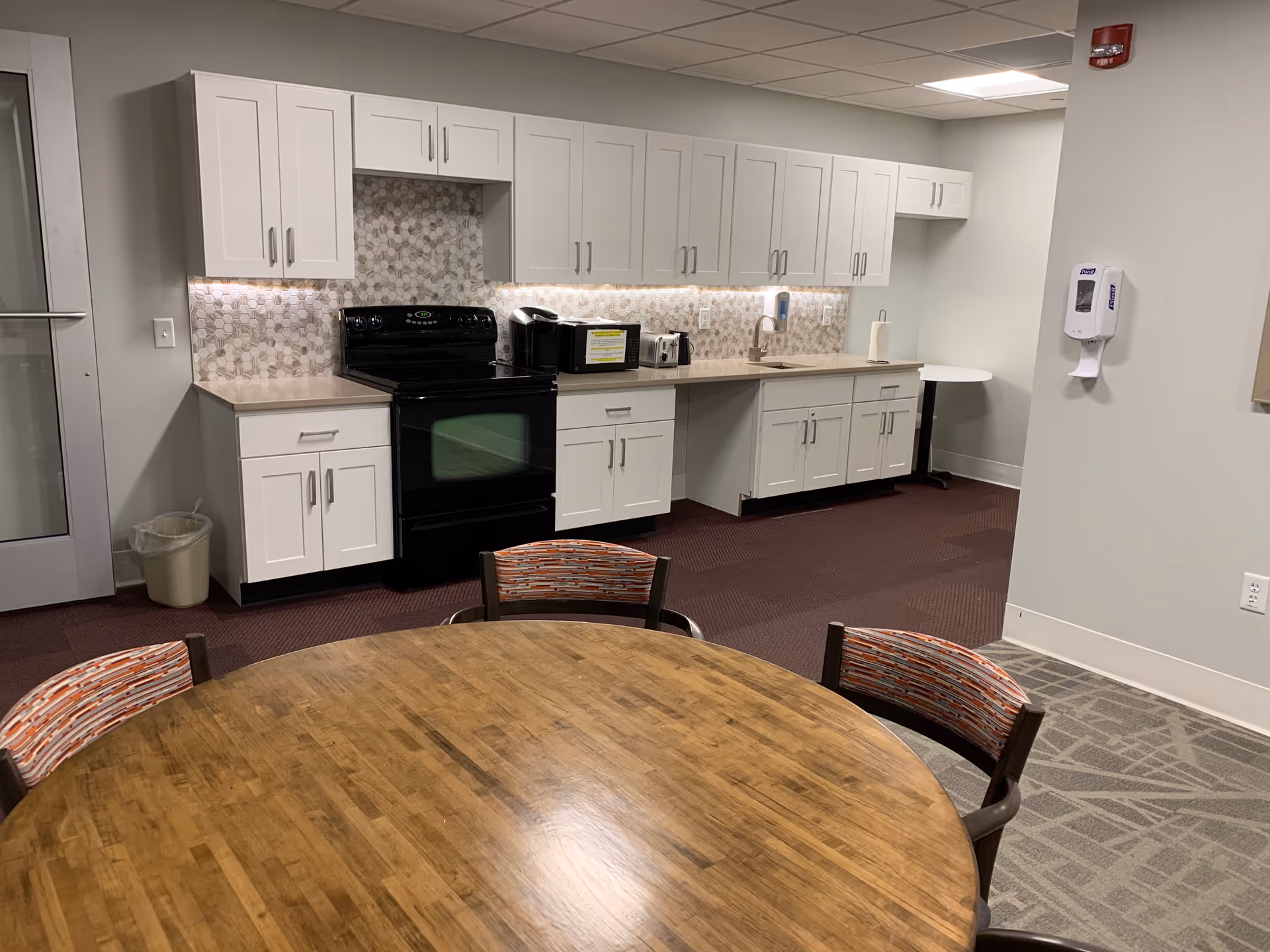 A clean and modern kitchen area with white cabinets, a black stove, a microwave, a toaster, and a sink. There is a round wooden table with four chairs featuring patterned cushions in the foreground. The floor has a combination of carpet and tile, and a hand sanitizer dispenser is mounted on the wall.
