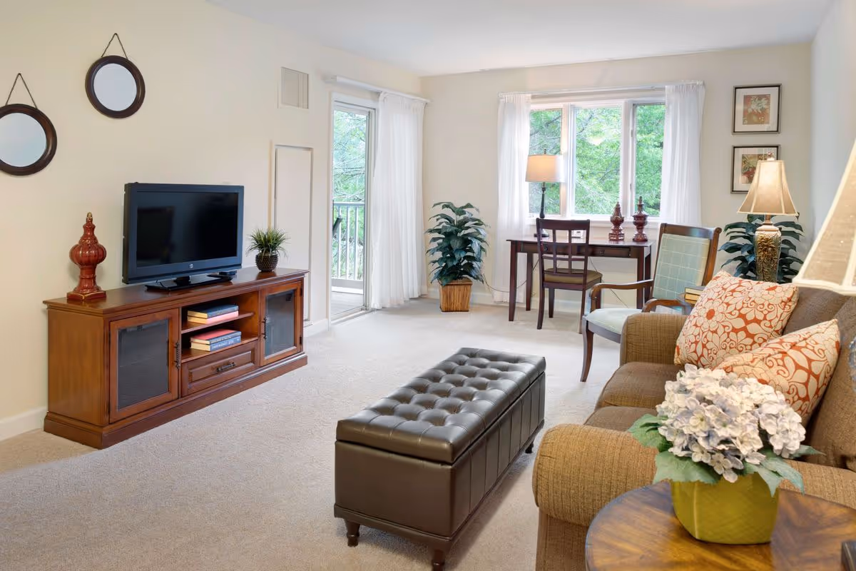 Bright furnished living room with a sofa, tufted ottoman, TV stand, and a desk by a large window.