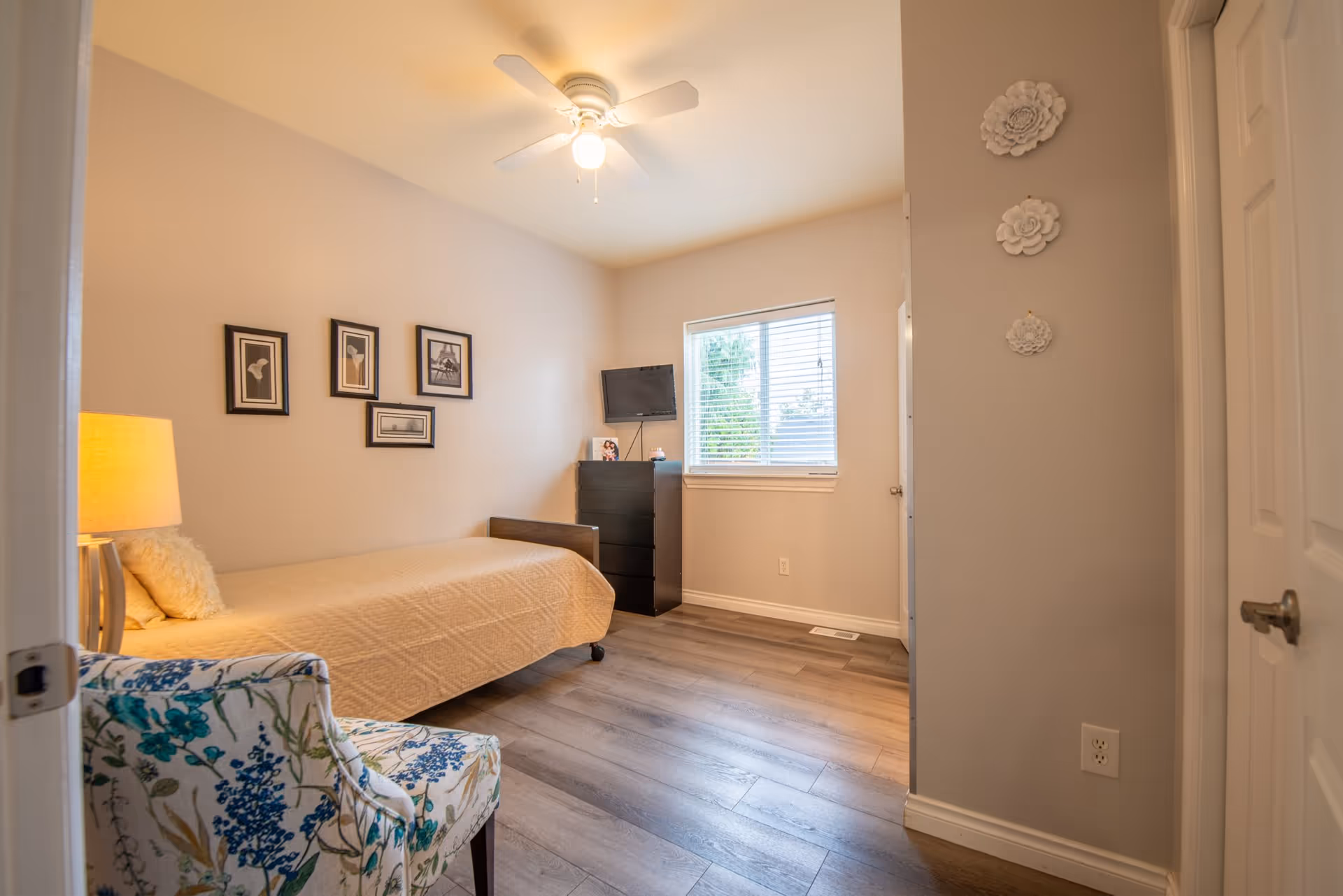 Tidy single bedroom with a bed, floral armchair, dresser with a small TV, window, wall art, and a ceiling fan.