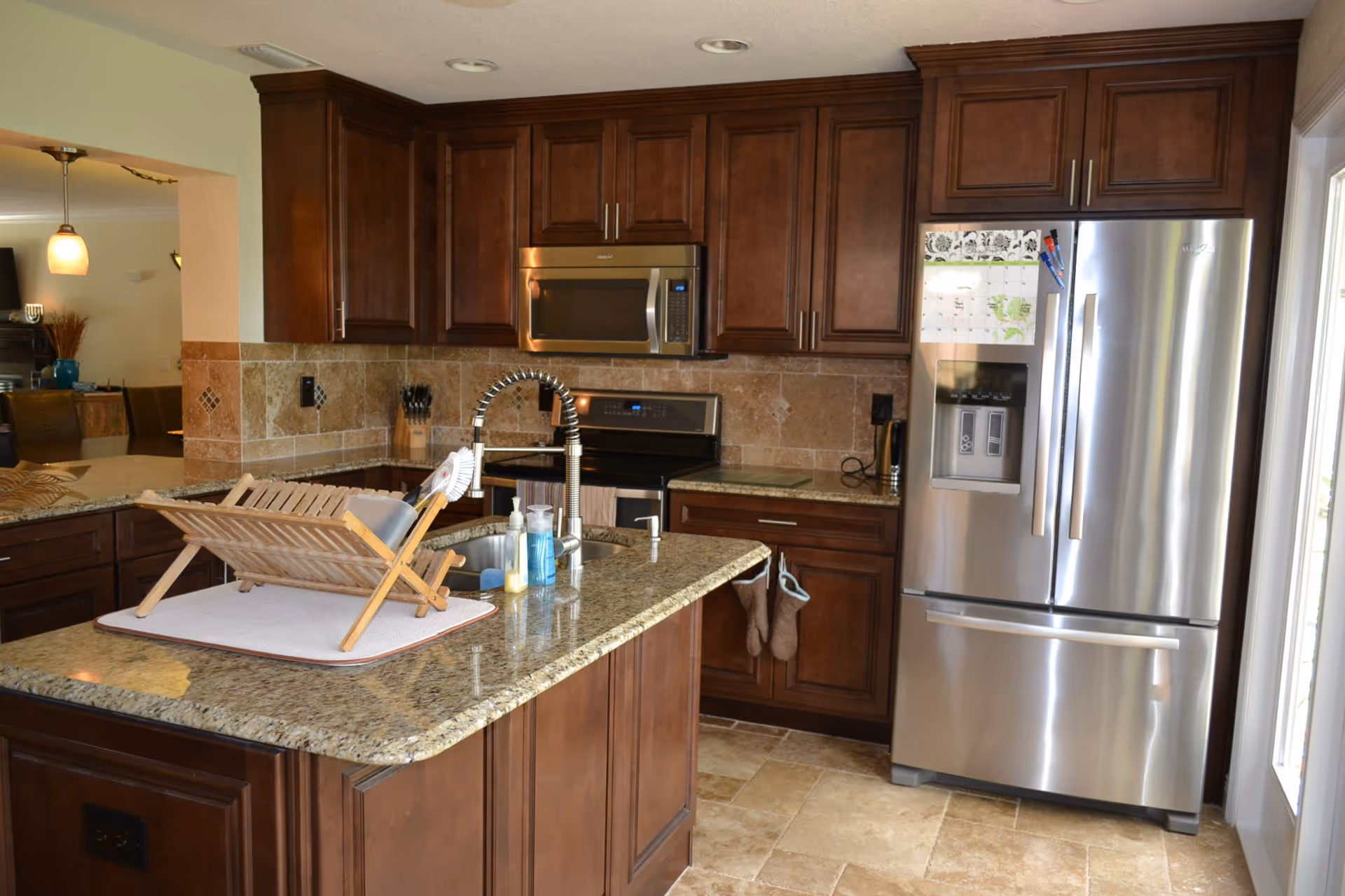 A modern kitchen with dark wooden cabinets, granite countertops, and a tiled backsplash. The kitchen features a stainless steel refrigerator with a calendar and magnets on the door, a built-in microwave above a stove, and a sink with a flexible spring faucet on the island. There is a wooden dish rack on the island countertop, along with soap dispensers and a dish brush. The kitchen opens into a dining or living area with visible chairs and pendant lighting.
