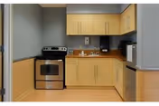 A small kitchen area with light wood cabinets, a stainless steel electric stove, a microwave, a sink, and a paper towel dispenser mounted on the wall. The walls are painted gray with a wooden wainscot paneling.