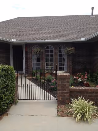 Front exterior view of a brick house with a black metal gate, a concrete pathway leading to a black front door, three arched windows with white curtains, hanging flower baskets, and landscaped garden beds with various plants and flowers.