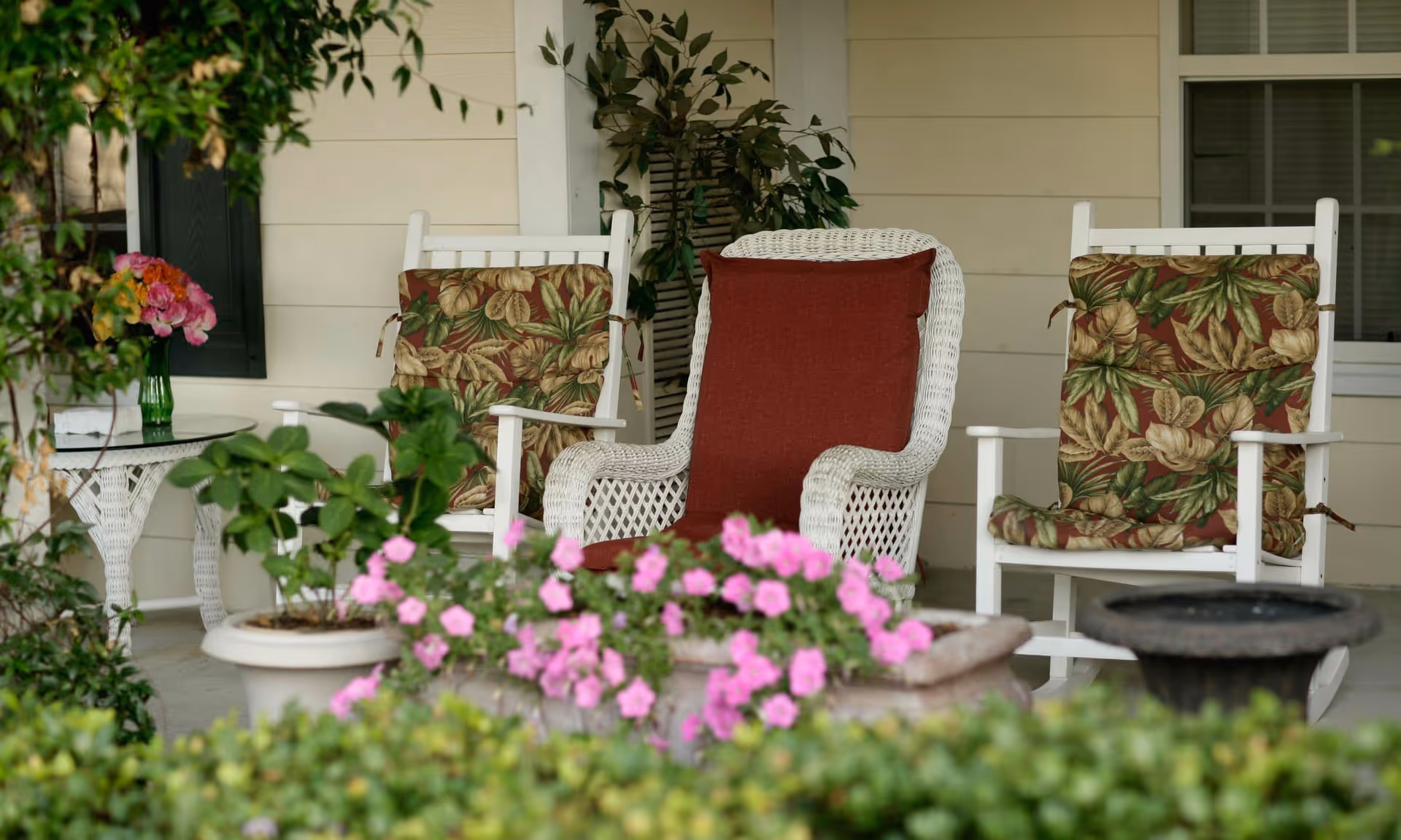 A cozy outdoor porch area with three chairs, two white wooden rocking chairs with floral cushions and one white wicker chair with a red cushion. There are potted plants and pink flowers in the foreground, a small white wicker side table with a vase of flowers, and greenery around the porch.