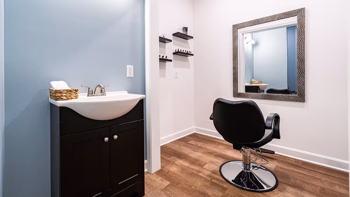 A small salon area with a black salon chair facing a large decorative mirror mounted on a white wall. To the left, there is a sink with a black cabinet below and a basket with towels on top. The wall behind the sink is painted light blue, and there are three small black shelves on the adjacent white wall holding various nail polish bottles. The floor is wood with a warm tone.