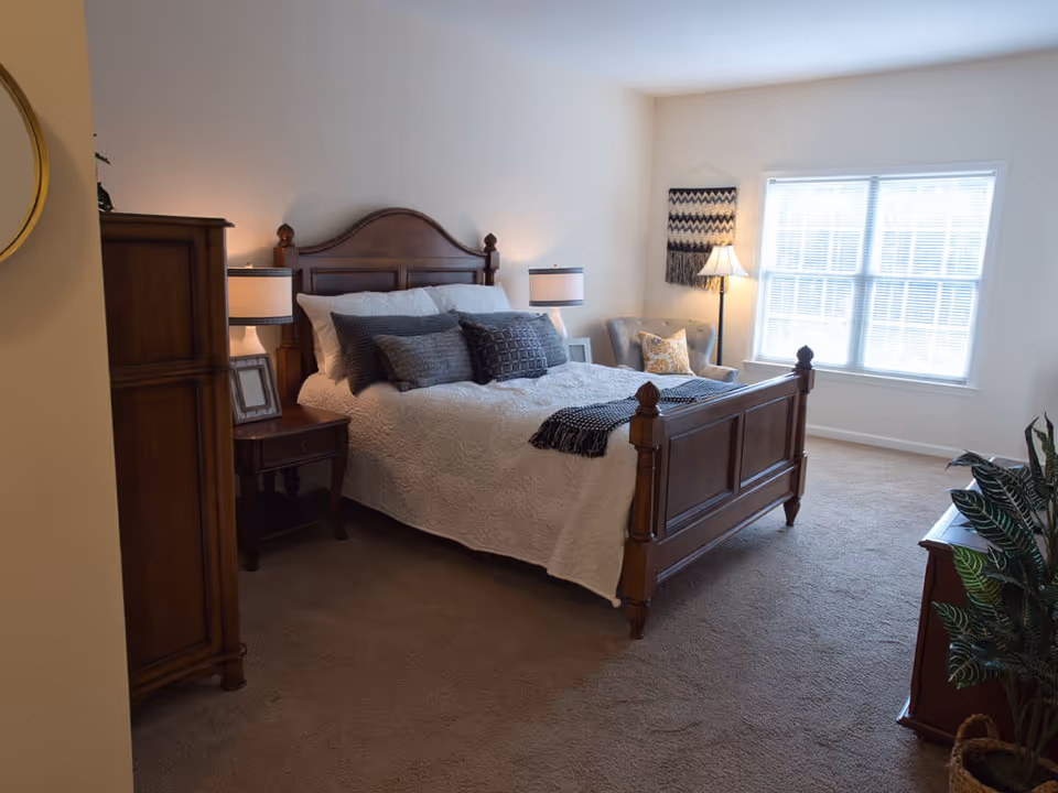 A cozy bedroom with a wooden bed frame, white bedding, and multiple pillows. There are two bedside tables with lamps, a wooden dresser, a comfortable armchair near a large window with blinds, a floor lamp, and a decorative wall hanging. The room has beige carpeting and neutral-colored walls.