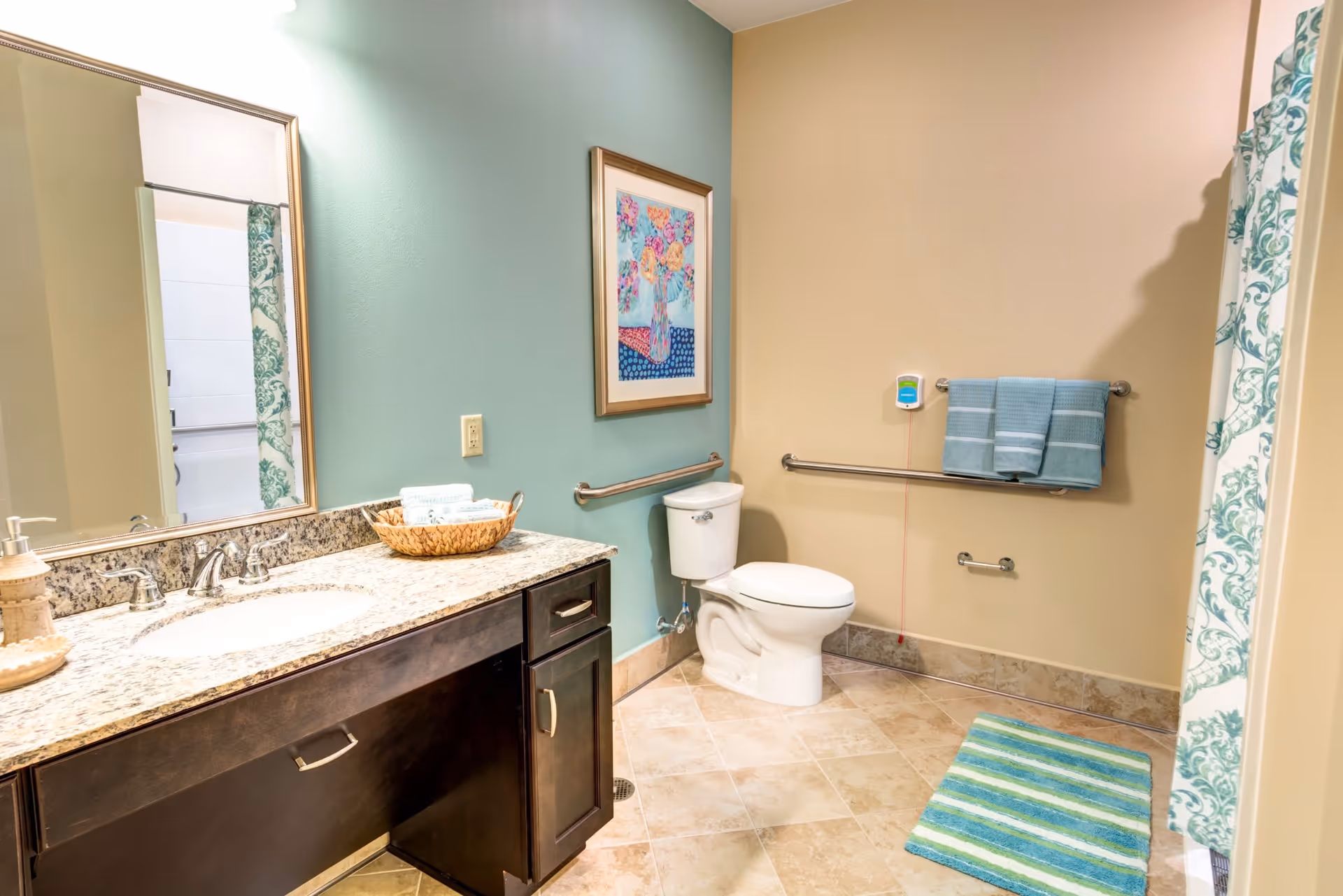 A clean and spacious bathroom with a granite countertop vanity featuring a sink and faucet, a large mirror above the vanity, a toilet with grab bars on the walls, a towel rack with blue towels, a colorful framed artwork on the wall, a shower curtain with a green and white pattern, and a striped blue and green bath mat on the tiled floor.