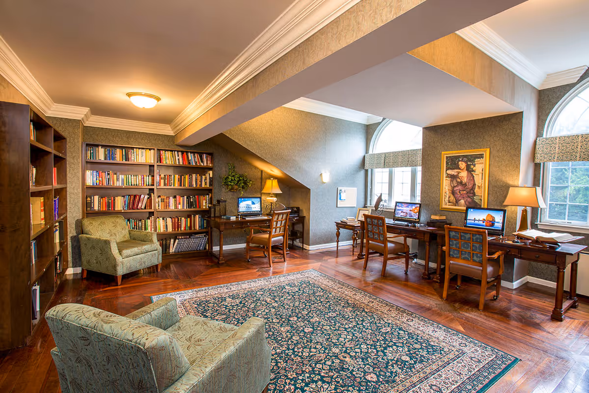 A cozy library and computer area in a senior living facility with wooden bookshelves filled with books, two upholstered armchairs, three wooden desks each with a computer and chair, a patterned area rug on polished hardwood floors, and large arched windows letting in natural light.