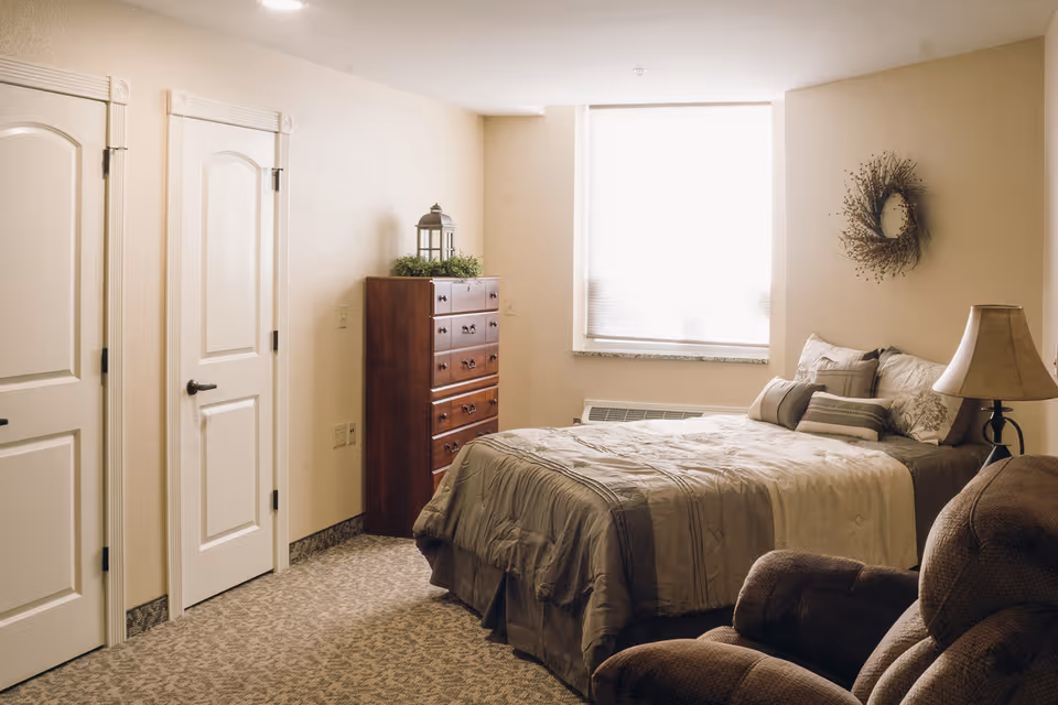Cozy furnished bedroom with a bed, wooden dresser, armchair and lamp under a bright window.