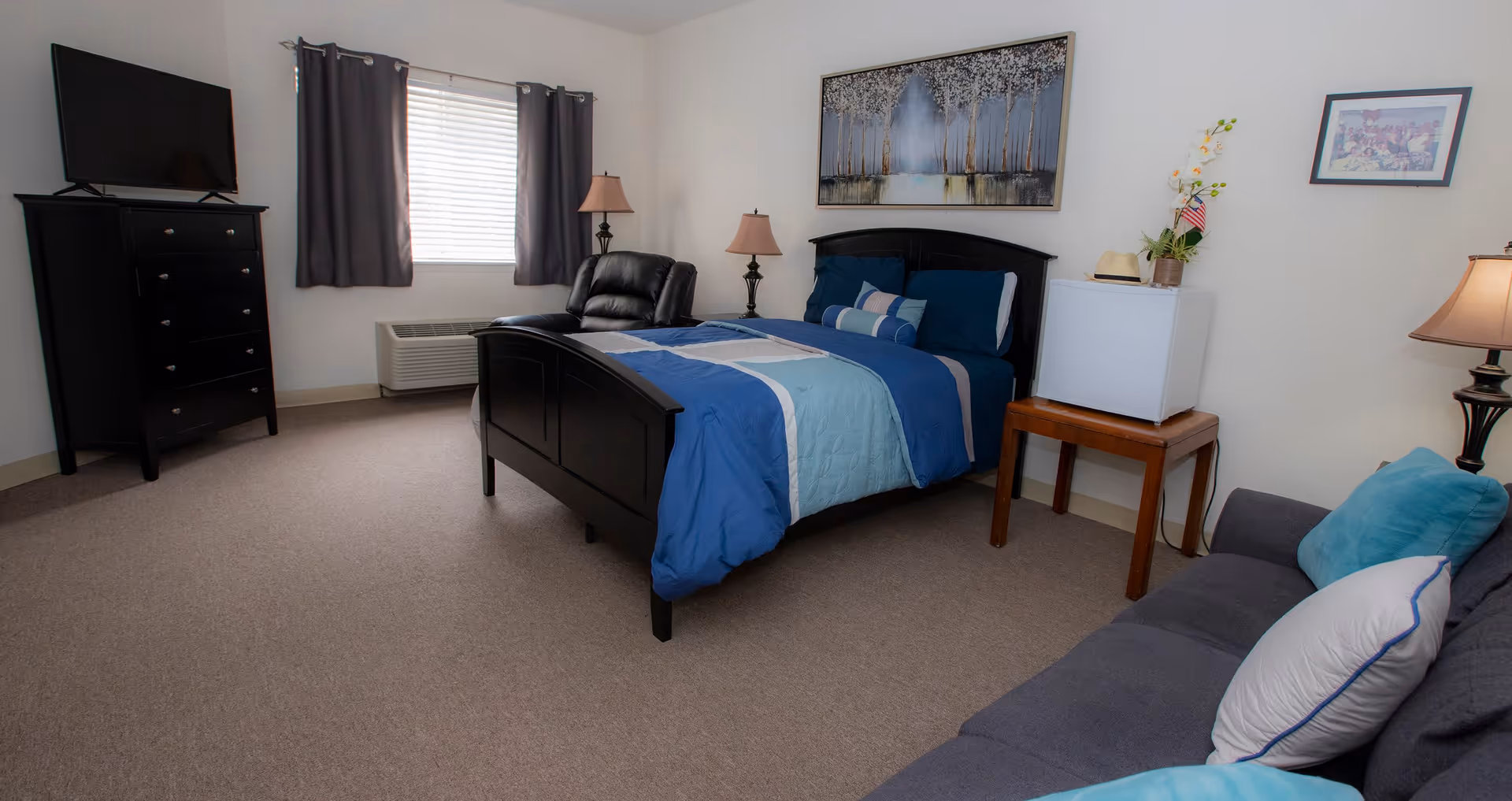 A bedroom in Colonial Assisted Living at Palm Beach featuring a bed with blue and light blue bedding, a black leather recliner, a black dresser with a flat-screen TV on top, a window with dark curtains, two lamps on either side of the bed, a small white refrigerator on a wooden table, a gray couch with blue and white pillows, and wall art above the bed and on the adjacent wall.