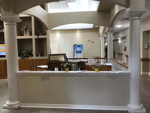 Interior view of a senior living facility reception area with a white counter in the foreground, two white columns on either side, potted plants on the counter, wooden cabinets and shelves with plants and decorations behind the counter, and a hallway extending to the right.