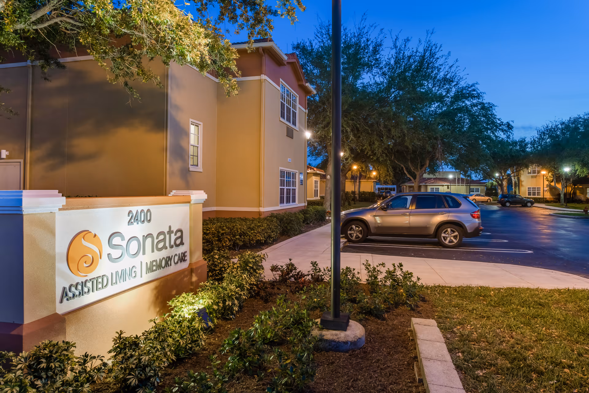 Exterior view of Sonata Assisted Living and Memory Care facility at dusk, showing the building, illuminated sign with the address 2400, a parking lot with cars, trees, and a clear blue sky.