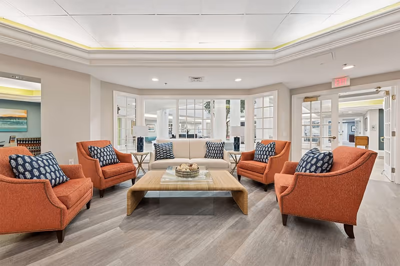 A bright and spacious living room area with a beige sofa and four orange armchairs arranged around a wooden coffee table. The room features large windows and glass doors, light-colored walls, and a tray ceiling with recessed lighting. Decorative pillows on the chairs and sofa add a touch of pattern and color.