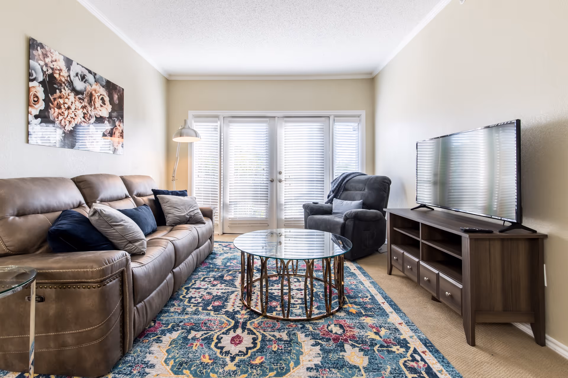 Bright living room with a leather sofa, recliner, glass coffee table, TV on a wooden console, and a patterned rug in front of French doors.