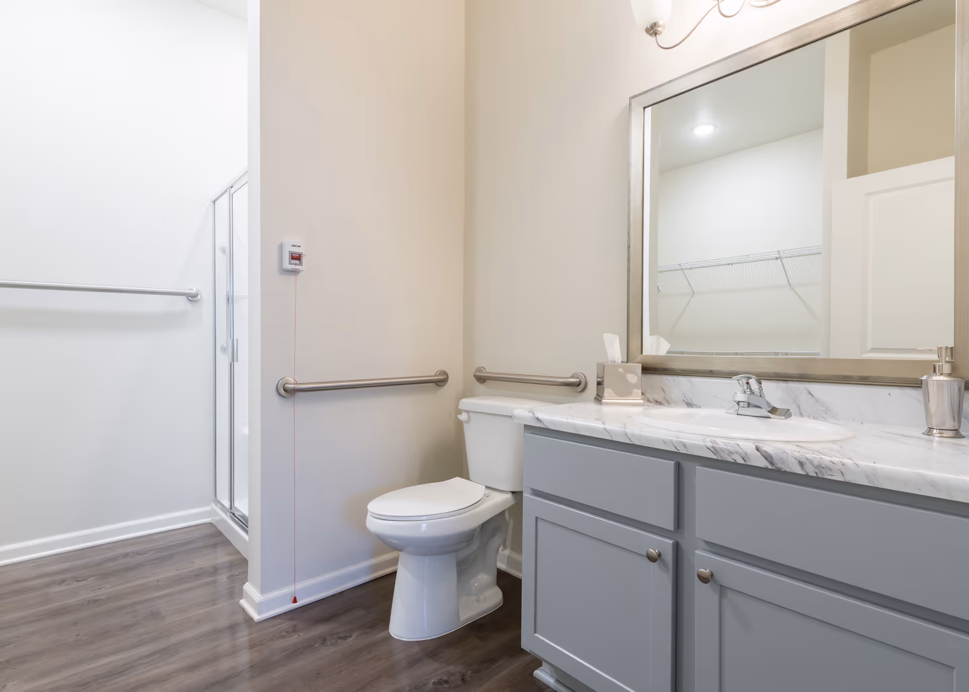 A clean and modern bathroom with a white toilet, a gray vanity with a marble countertop, a large mirror above the sink, and a glass-enclosed shower. The bathroom has light-colored walls, wood-style flooring, and safety grab bars near the toilet and shower area.