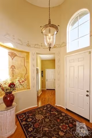 A bright entryway with a white front door and a large arched window above it. The space features a hanging glass light fixture, a decorative floral rug on wooden flooring, a small round table with a red vase holding flowers, and a wall with a framed mural painting. The walls are painted in a light color with subtle decorative stenciling near the ceiling.