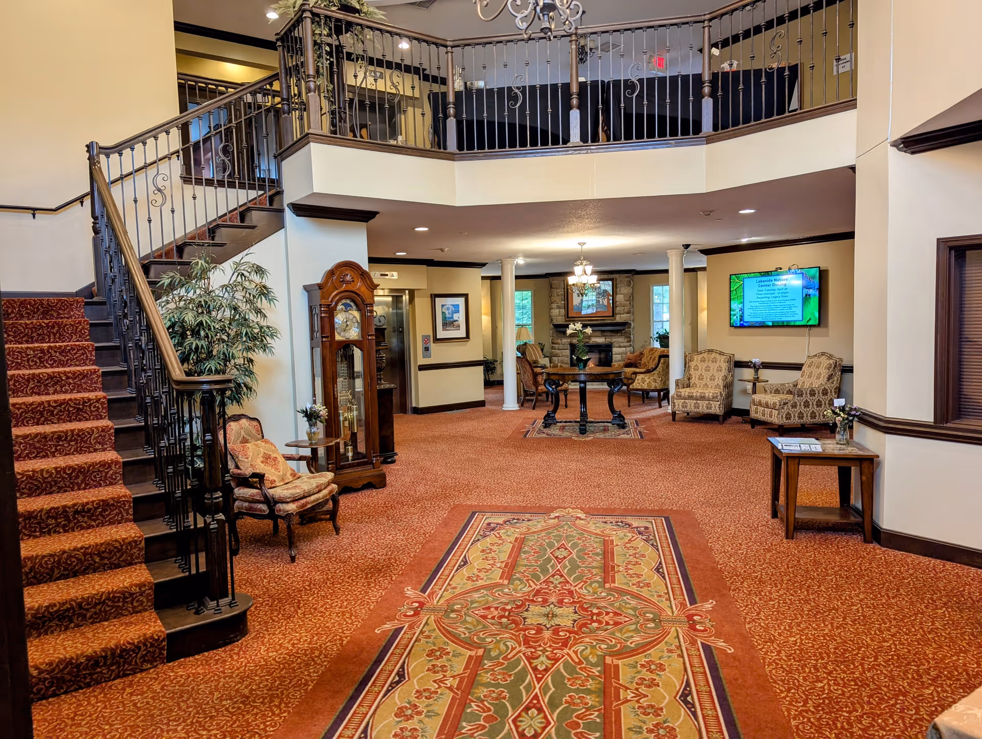 Interior view of a senior living community common area with a staircase on the left, a grandfather clock, upholstered chairs, a decorative rug, and a seating area with armchairs and a stone fireplace in the background. A television screen is mounted on the wall to the right.
