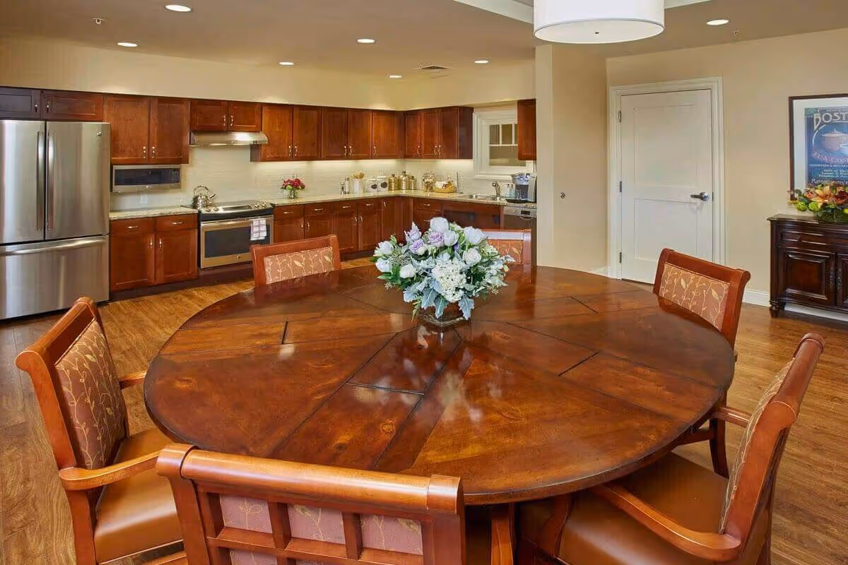A spacious kitchen and dining area with wooden cabinets, stainless steel refrigerator, stove, and dishwasher. In the foreground, there is a large round wooden dining table with six cushioned chairs and a floral centerpiece. The room has warm lighting and a wooden floor.